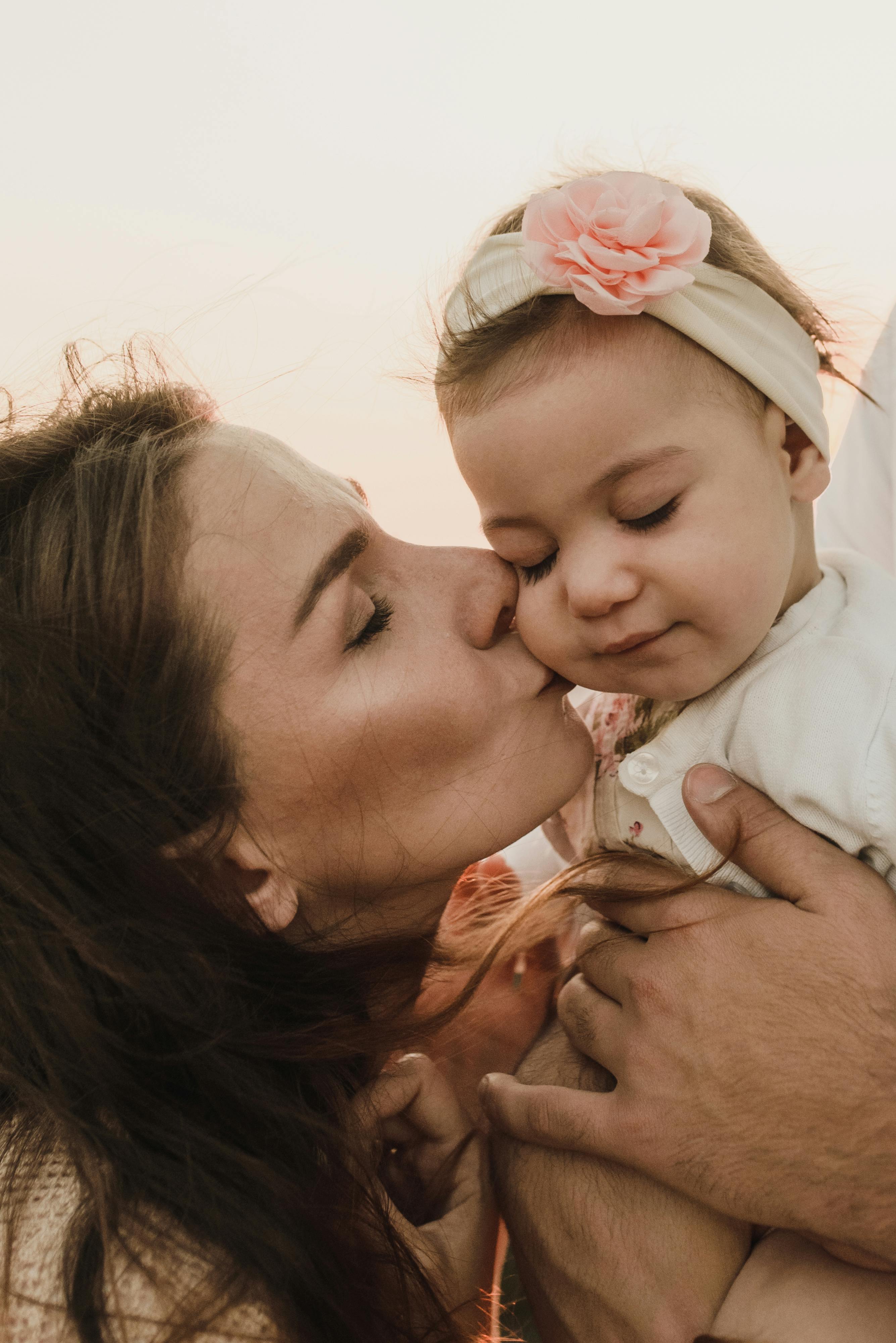 Tender moment of a mother kissing her baby daughter outdoors at sunset, showcasing love and family bond.